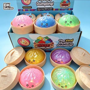 May include: A display box of Mystery Dumpling toys featuring glittery, squishy, round dumpling figures with smiling faces, each housed in a small, tan, circular bamboo-style steamer container. The box includes text reading The Viral Mystery Dumpling.