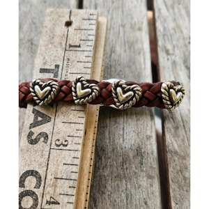 May include: Braided brown leather bracelet with silver-toned heart-shaped accents. The bracelet is shown next to a wooden ruler. The ruler is marked with inches.