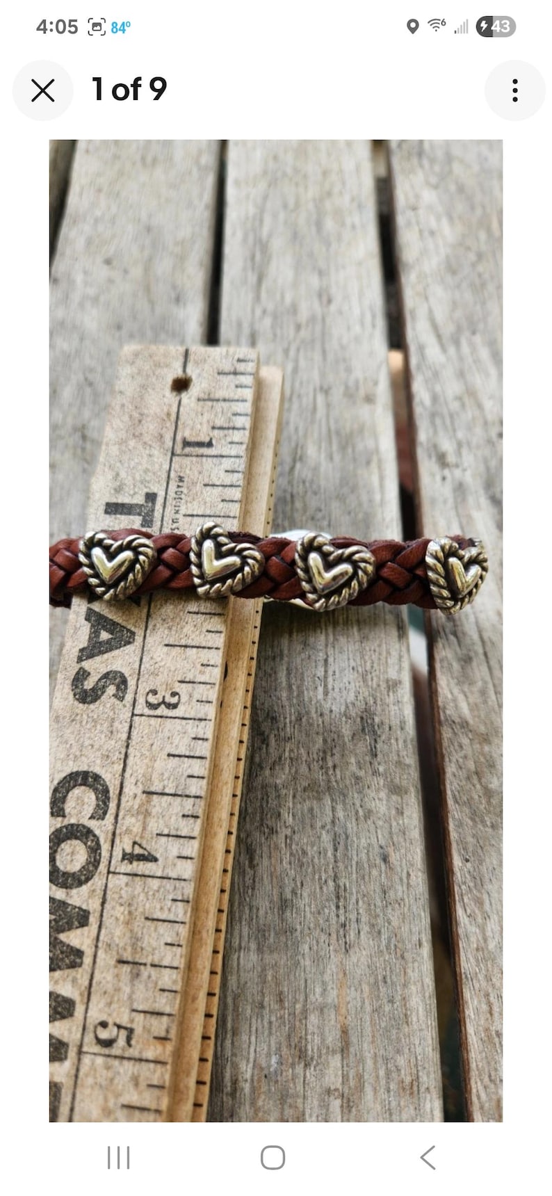 May include: Braided brown leather bracelet with silver-toned heart-shaped accents. The bracelet is shown next to a wooden ruler. The ruler is marked with inches.