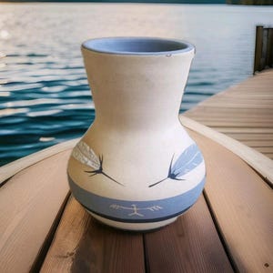 May include: A cream-colored ceramic vase with a flared rim and a rounded body. The vase features two painted feathers and a horizontal blue band near the base, which contains a small, white, stylized human figure graphic centered on the front.