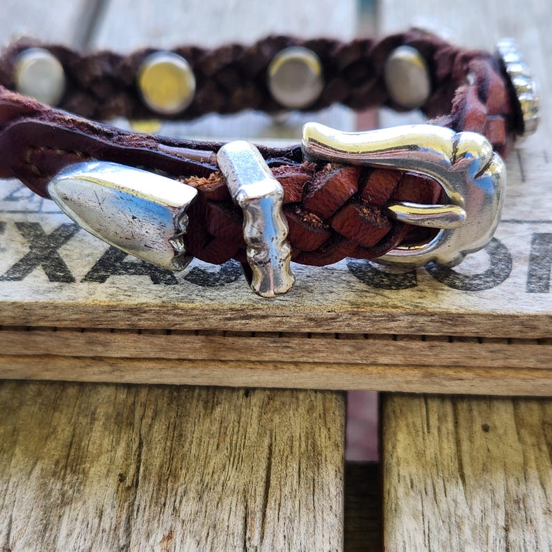 May include: A brown leather bracelet with a braided design and silver-colored metal accents. The bracelet features a buckle closure and decorative studs. The leather has a rich, dark brown color.