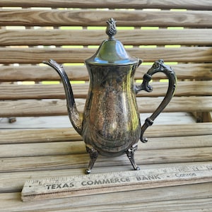 May include: A silver-plated coffee pot with a tapered body, a curved spout, and an ornate handle. It features a removable lid with a finial and stands on three decorative feet. A wooden ruler shows the pot is approximately 9 inches tall.