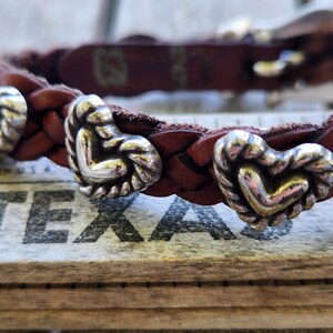May include: Braided brown leather bracelet with silver heart-shaped accents. The bracelet features a woven design and decorative metal hearts. The bracelet is resting on a wooden surface with the word "TEXAS" visible.