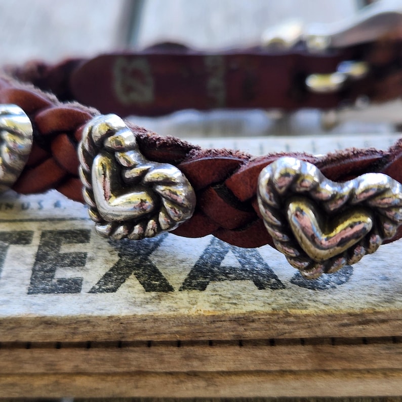 May include: Braided brown leather bracelet with silver heart-shaped accents. The bracelet features a woven design and decorative metal hearts. The bracelet is resting on a wooden surface with the word "TEXAS" visible.