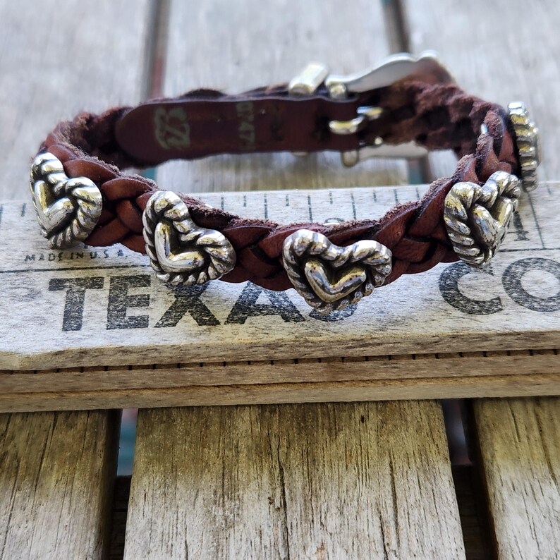 May include: Braided brown leather bracelet with silver heart-shaped accents. The bracelet features a silver clasp and is displayed on a wooden surface with the word "TEXAS" visible.