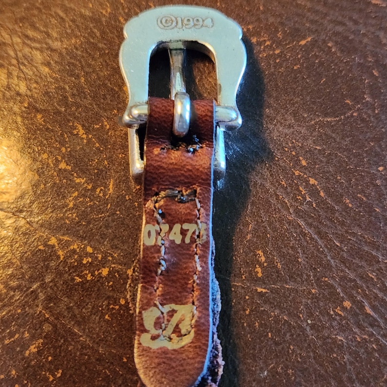May include: Close-up of a brown leather belt buckle with a silver-colored metal buckle. The leather strap is stamped with the numbers "047" and the letters "SZ". The buckle is stamped with "&copy;1994".