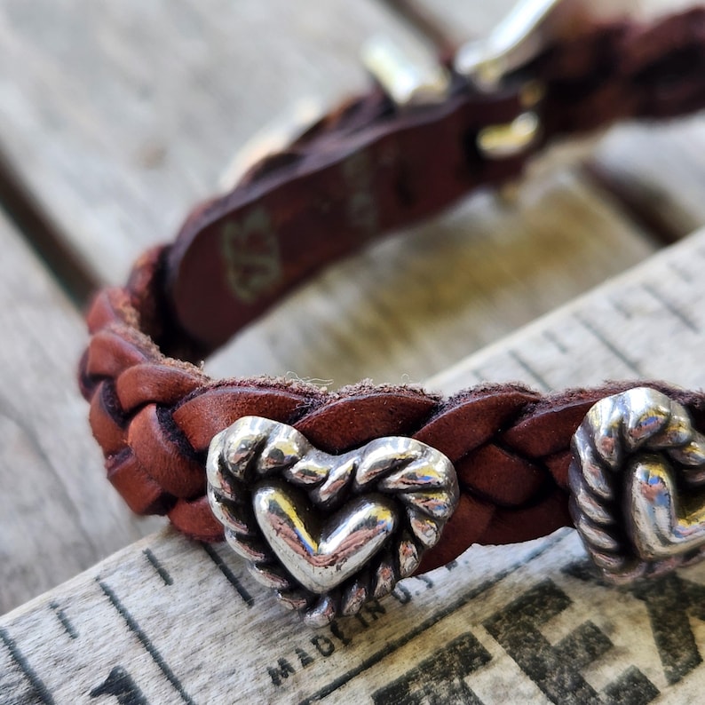 May include: Braided brown leather bracelet with silver heart-shaped accents. The bracelet features a woven design and a silver buckle. The heart-shaped accents are detailed with a rope-like border. The bracelet is on a wooden surface.