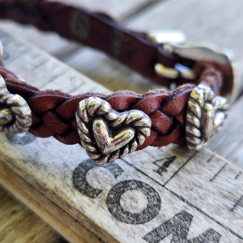 May include: Braided brown leather bracelet with silver heart-shaped charms. The bracelet features a silver buckle and is displayed on a wooden surface. A close-up shot highlights the texture and details of the jewelry.