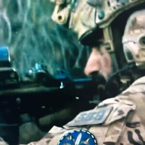 Puede incluir: Una persona con uniforme de camuflaje color canela y un parche circular azul con un emblema de espada blanca en la manga. El individuo lleva un casco t&aacute;ctico con equipo montado y sostiene un rifle negro en posici&oacute;n de enfoque frontal.