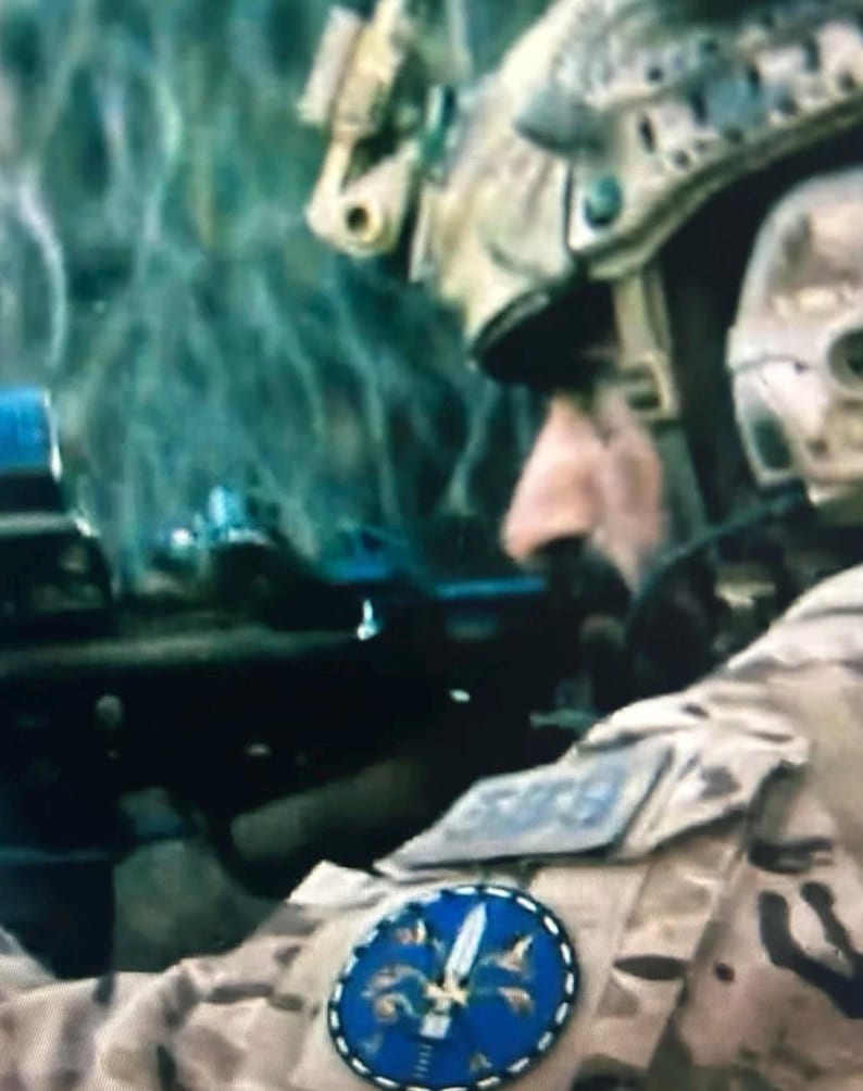Puede incluir: Una persona con uniforme de camuflaje color canela y un parche circular azul con un emblema de espada blanca en la manga. El individuo lleva un casco t&aacute;ctico con equipo montado y sostiene un rifle negro en posici&oacute;n de enfoque frontal.