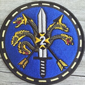 Puede incluir: Un parche bordado circular con fondo azul y una espada plateada central apuntando hacia arriba. Un n&uacute;mero 2 amarillo est&aacute; centrado en la espada. Un drag&oacute;n de varias cabezas con escamas amarillas y negras se enrosca alrededor de la espada y dos flechas.