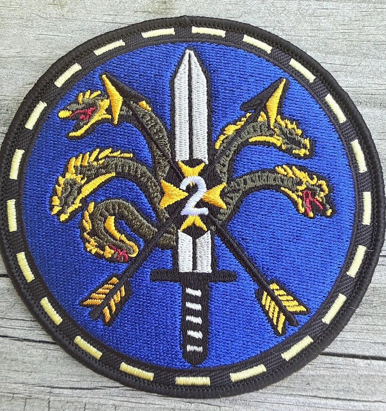 Puede incluir: Un parche bordado circular con fondo azul y una espada plateada central apuntando hacia arriba. Un n&uacute;mero 2 amarillo est&aacute; centrado en la espada. Un drag&oacute;n de varias cabezas con escamas amarillas y negras se enrosca alrededor de la espada y dos flechas.