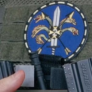 Puede incluir: Un parche circular con fondo azul, una espada plateada central, dos flechas cruzadas y una criatura hidra de varias cabezas. Una cruz blanca con el n&uacute;mero 2 est&aacute; centrada en la espada. Debajo, un bol&iacute;grafo t&aacute;ctico negro y un cargador Surefire.