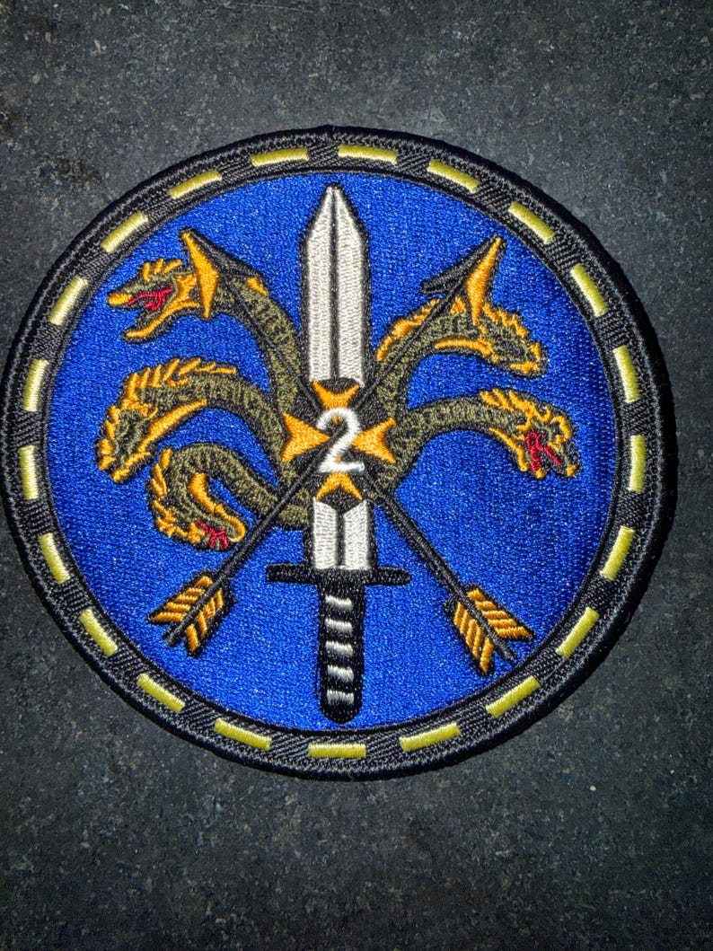 Puede incluir: Un parche bordado circular con fondo azul y una espada plateada central apuntando hacia arriba. Un drag&oacute;n dorado de varias cabezas se enrosca alrededor de la espada, cruzada por dos flechas doradas. El n&uacute;mero 2 est&aacute; en el centro, borde negro con rayas amarillas.