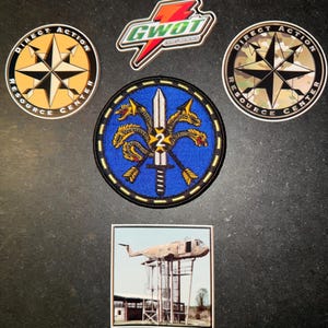 May include: A collection of patches and a sticker featuring two circular Direct Action Resource Center emblems, a GWOT Thirst Quencher lightning bolt sticker, a blue circular patch with a hydra and sword, and a square sticker of a helicopter.