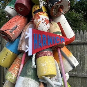 May include: A red felt pennant with the word HARWICH in blue block letters, featuring a white vertical strip with two white tabs on the left side, displayed against a cluster of weathered, multi-colored cylindrical nautical lobster buoys.