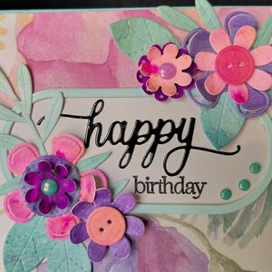 Può includere: Un biglietto d'auguri fatto a mano con uno sfondo ad acquerello nei toni del rosa e del viola. Il biglietto è decorato con fiori di carta stratificati in rosa, viola e verde acqua, accentuati da bottoni. Il testo recita happy birthday in nero.