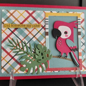 May include: A rectangular greeting card with a red border, featuring a plaid background in blue, yellow, and red. A die-cut pink parrot sits on a branch inside a frame. Green paper monstera leaves and a yellow label with the text Did someone say cake? are attached.