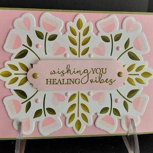 May include: A rectangular greeting card with a light pink background featuring a white die-cut overlay decorated with pink tulips, olive green leaves, and small pink dots. A central pink label displays the gold embossed text wishing YOU HEALING vibes.