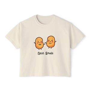 May include: A cream-coloured t-shirt with a cartoon illustration of two smiling potato characters wearing bow ties. The text "Best Spuds" is printed below the potatoes in a black, handwritten-style font. The shirt has short sleeves and a crew neck.