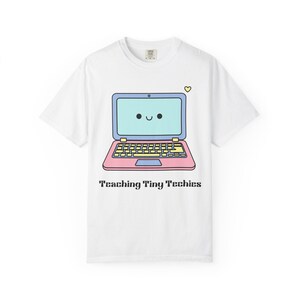 Peut inclure: T-shirt blanc avec un ordinateur portable de dessin animé. L'ordinateur portable a un visage souriant sur l'écran et est rose, bleu et jaune. Le texte "Teaching Tiny Techies" est imprimé en dessous.