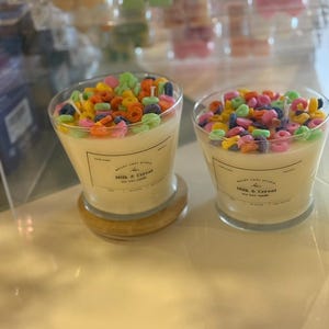 May include: Two glass candles with a creamy wax base, topped with colorful, cereal-shaped wax pieces. The candles are labeled "Milk & Cereal" and sit on a light-colored surface. The background shows other candles in clear containers.