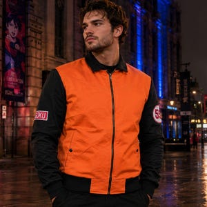 Pode incluir: Um blusão bomber com fecho de correr, tronco cor de laranja brilhante e mangas pretas contrastantes. O casaco inclui uma gola preta, punhos canelados pretos e dois emblemas: um emblema retangular rosa com o texto GOKU e um circular branco com 59.
