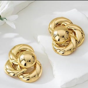 May include: A pair of gold-toned earrings featuring a polished, reflective finish. Each earring consists of a central smooth sphere surrounded by three interconnected, twisted, rope-like circular loops, creating a dimensional, layered design.