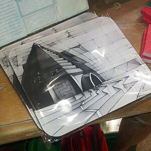 May include: A stack of square coasters with a black and white architectural design. The coasters are packaged in clear plastic. Other items on the table include a red notebook, a box of butterflies, and a notepad.