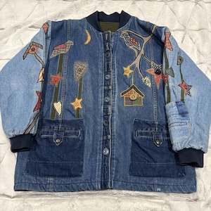May include: A denim jacket with a variety of colorful, whimsical appliqués. The jacket features a dark blue ribbed collar and cuffs, two front pockets, and a button-up closure. Appliqués include birds, stars, hearts, and houses.
