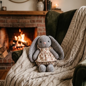 May include: A gray crocheted plush rabbit toy with long ears, black safety eyes, and a small pink stitched nose. The rabbit wears a cream-colored dress and matching bloomers featuring a delicate red and green floral pattern.