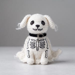 Puede incluir: Una figura de perro tejida a ganchillo en color blanco con detalles de caja torácica esquelética bordados en negro en el pecho y una boca cosida. El perro lleva un collar negro con un pequeño dije de calavera plateado, ojos negros y una base.