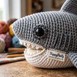 May include: A grey crocheted shark plush toy with a white underbelly and sharp white teeth. The shark features a black plastic eye and a small fabric tag attached to the side that reads TrippyPeelz HANDMADE | CRAFTED WITH CARE in blue text.