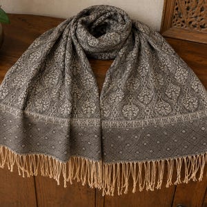 May include: A rectangular scarf featuring a dark gray and beige intricate floral damask pattern across the main body, with a geometric diamond and dot border design at the ends, finished with long, light beige fringe tassels along the bottom edges.