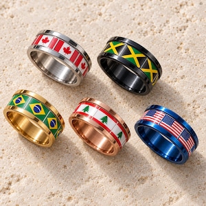 May include: Five metal rings featuring national flag designs. One silver ring with the Canadian maple leaf, a black ring with the Jamaican flag, a gold ring with the Brazilian flag, a rose gold ring with the Lebanese cedar, and a blue ring with the US flag.