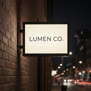 Può includere: Un'insegna rettangolare illuminata montata a parete con una cornice nera fissata su un muro di mattoni rossi. L'insegna presenta una superficie bianca luminosa con il testo "LUMEN CO." stampato in un carattere nero sans-serif centrato.