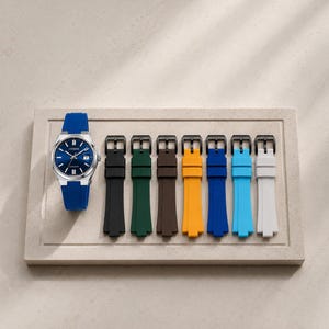 Könnte beinhalten: Eine silberfarbene Uhr mit blauem Zifferblatt und blauem Silikonarmband wird neben einer Auswahl an bunten Uhrenarmbändern präsentiert. Die Armbänder sind schwarz, grün, braun, gelb, blau, hellblau und weiß. Die Uhr und die Armbänder befinden sich auf einem rechteckigen, hellen Tablett.