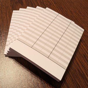 May include: A stack of lined notepads with a light gray and white striped pattern. The notepads have a white border at the bottom and are arranged on a dark wooden surface. The paper appears to be blank.