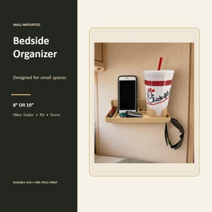 May include: A beige wall-mounted bedside organizer featuring a rectangular tray with a built-in cup holder. The organizer holds a smartphone, a set of keys, a remote control, and a large white beverage cup with a red straw and logo.