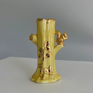 May include: A pale yellow ceramic vase shaped like a tree trunk with a gold-painted rim. The vase features a small gold squirrel figure perched on a branch and gold-painted leaf details on the front, with a textured bark-like surface finish.