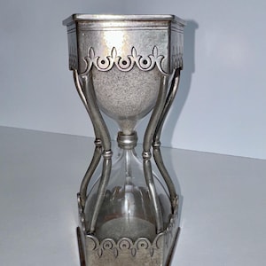 May include: An hourglass featuring a clear glass bulb containing grey sand, encased in a metallic silver frame. The frame has a hexagonal base and top with decorative cut-out arches and vertical supports connecting the two ends of the timer.