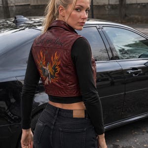 May include: A dark red leather vest featuring a graphic of a figure surrounded by orange and yellow flames on the back, with the embroidered text "Let me live" above the design. The vest is worn over a black long-sleeve shirt and black jeans.