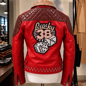 May include: A red leather jacket featuring brown quilted leather shoulders and a tan collar. The back displays a large embroidered patch with the text Lucky 38, a roulette wheel, and two white dice. The hem is accented with silver metal studs.