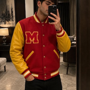 Pode incluir: Uma jaqueta varsity vermelha com mangas amarelas e uma letra M amarela no peito. A jaqueta possui fecho frontal com botões de pressão, dois bolsos com debrum amarelo e punhos e bainha canelados listrados nas cores vermelha, branca e amarela.