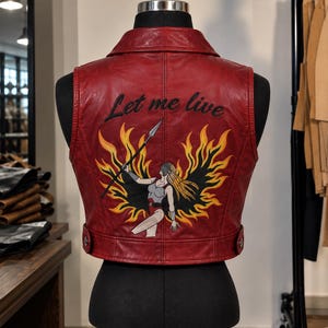 May include: A red leather vest featuring a back graphic of a warrior figure holding a spear amidst yellow and orange flames. The text "Let me live" is embroidered in black above the illustration. The vest has a collared neckline and side tabs.