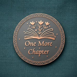 May include: A circular metal pin featuring a dark grey centre with rose gold accents. The design includes an open book with three flowers emerging from the pages and the text "One More Chapter" in a serif font, surrounded by a decorative border.