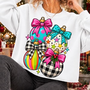 Può includere: Una felpa bianca a maniche lunghe con una stampa grafica di quattro decorazioni natalizie. Le decorazioni sono decorate con motivi rosa brillante, turchese e a quadri bianchi e neri, ognuna sormontata da un fiocco colorato e piccoli dettagli festivi.