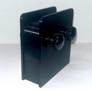 Peut inclure: Un dispositif de serrage noir, rectangulaire et imprimé en 3D, composé de deux plaques verticales parallèles reliées par un bloc central. Deux boutons de réglage noirs, circulaires et striés, sont situés sur le côté pour le serrage manuel.