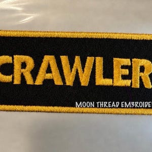 May include: A rectangular embroidered patch with a black background and a gold border. The word CRAWLER is stitched in large gold capital letters across the center. The text MOON THREAD EMBROIDERY is stitched in small white letters in the bottom right corner.