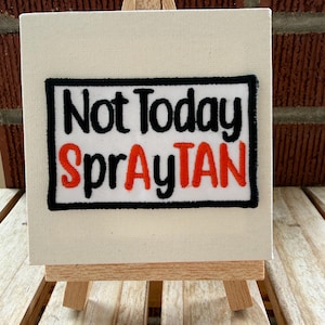 May include: A rectangular embroidered patch featuring the black and orange text Not Today SprayTAN on a white background, framed by a thick black border. The patch is mounted on a small, square white canvas displayed on a wooden easel.
